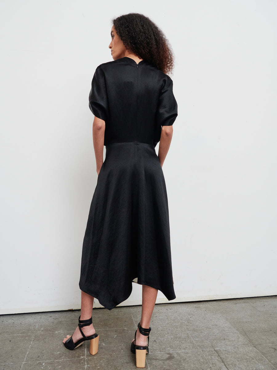 Someone with curly hair faces away from the camera, wearing the Patchwork Aki Wave Dress by Zero + Maria Cornejo. This black linen blend dress is distinguished by its puffed sleeves and asymmetrical hem. They pair it elegantly with black high-heeled sandals, standing on a concrete floor against a plain white wall.