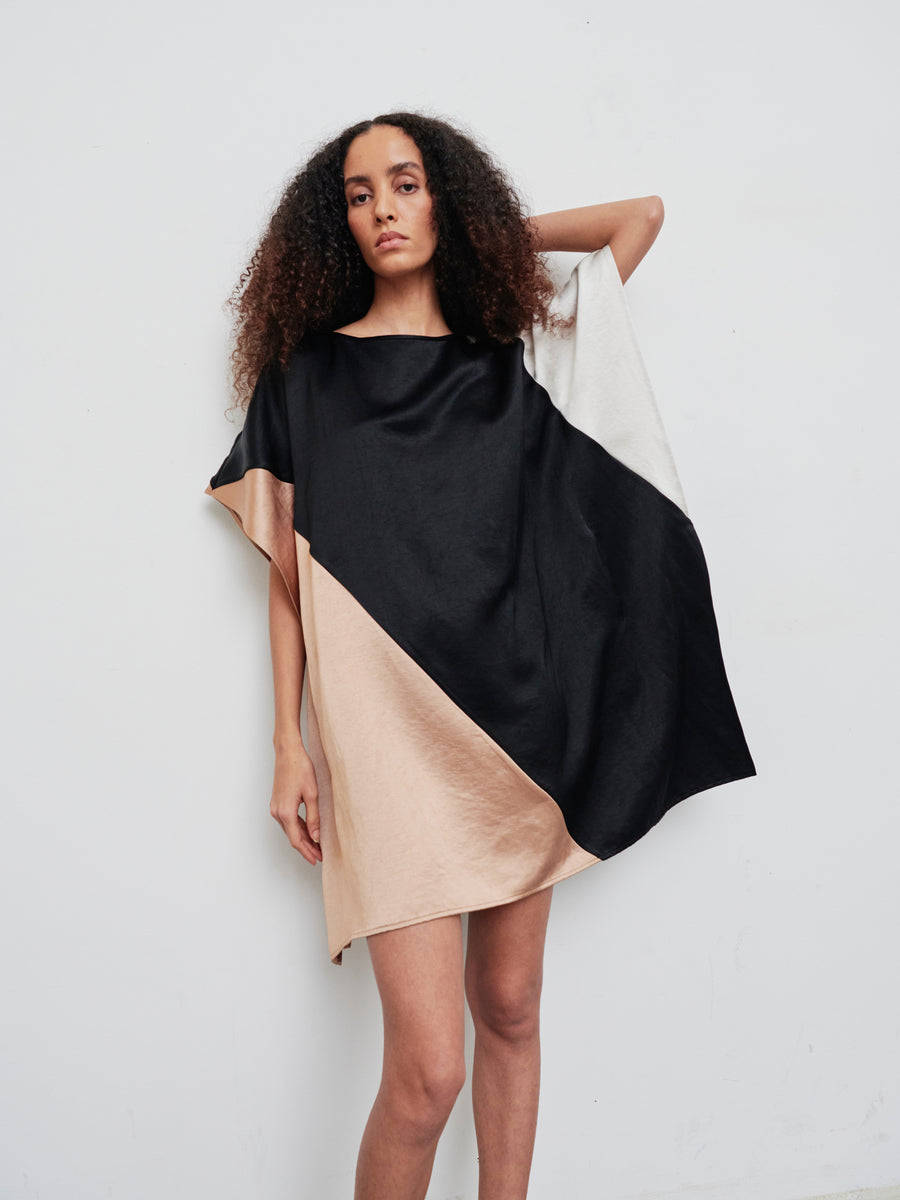 A person with curly hair is wearing the Dia Dress from Zero + Maria Cornejo, which features a knee-length, color-blocked design in black, beige, and white. They are standing against a plain white background. The linen-blend dress is loose-fitting and drapes elegantly over the shoulder.