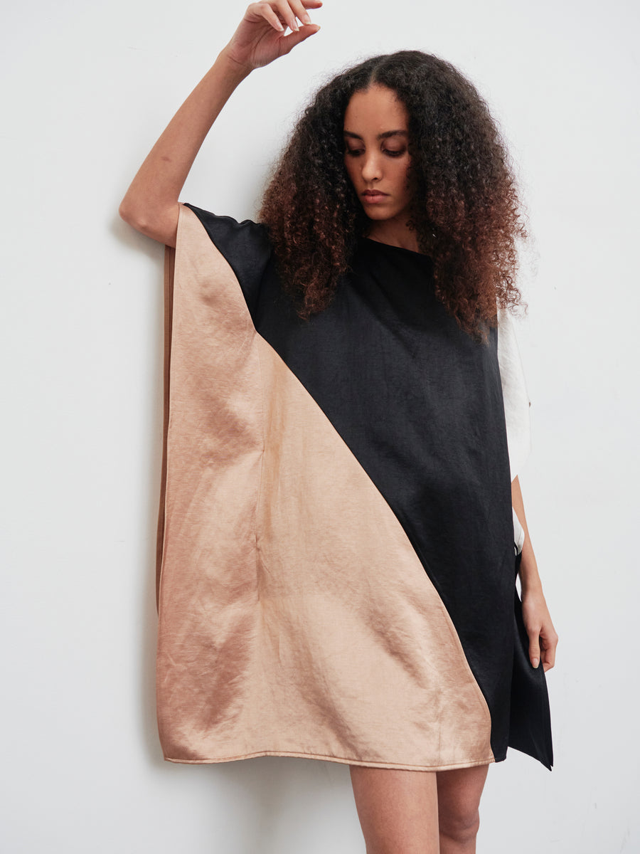 A person with curly hair wears the Dia Dress, a loose, knee-length color block outfit in black and beige by Zero + Maria Cornejo, crafted from a linen-blend. They stand against a plain white background, looking down with one arm slightly raised.