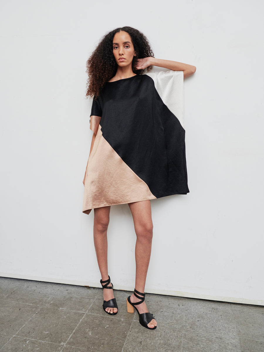 A person with curly hair stands against a white wall wearing the knee-length, short-sleeve Dia Dress by Zero + Maria Cornejo, which features black, beige, and white color blocks. They have one hand in their pocket and are wearing black strappy sandals with wooden heels.