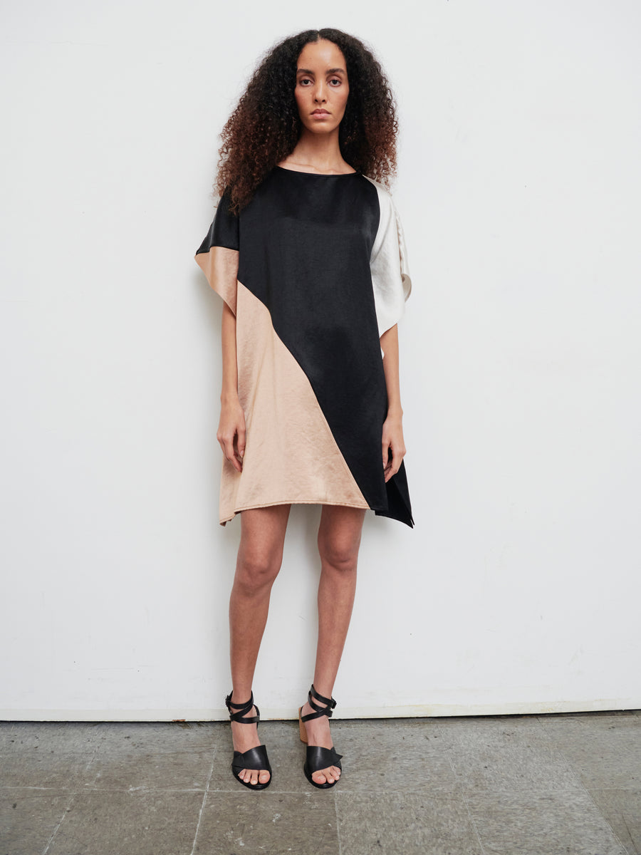 A woman stands against a plain white wall wearing the Dia Dress by Zero + Maria Cornejo, a knee-length linen-blend design featuring geometric sections in black, beige, and white. She has long, curly hair and pairs the dress with black strappy sandals.