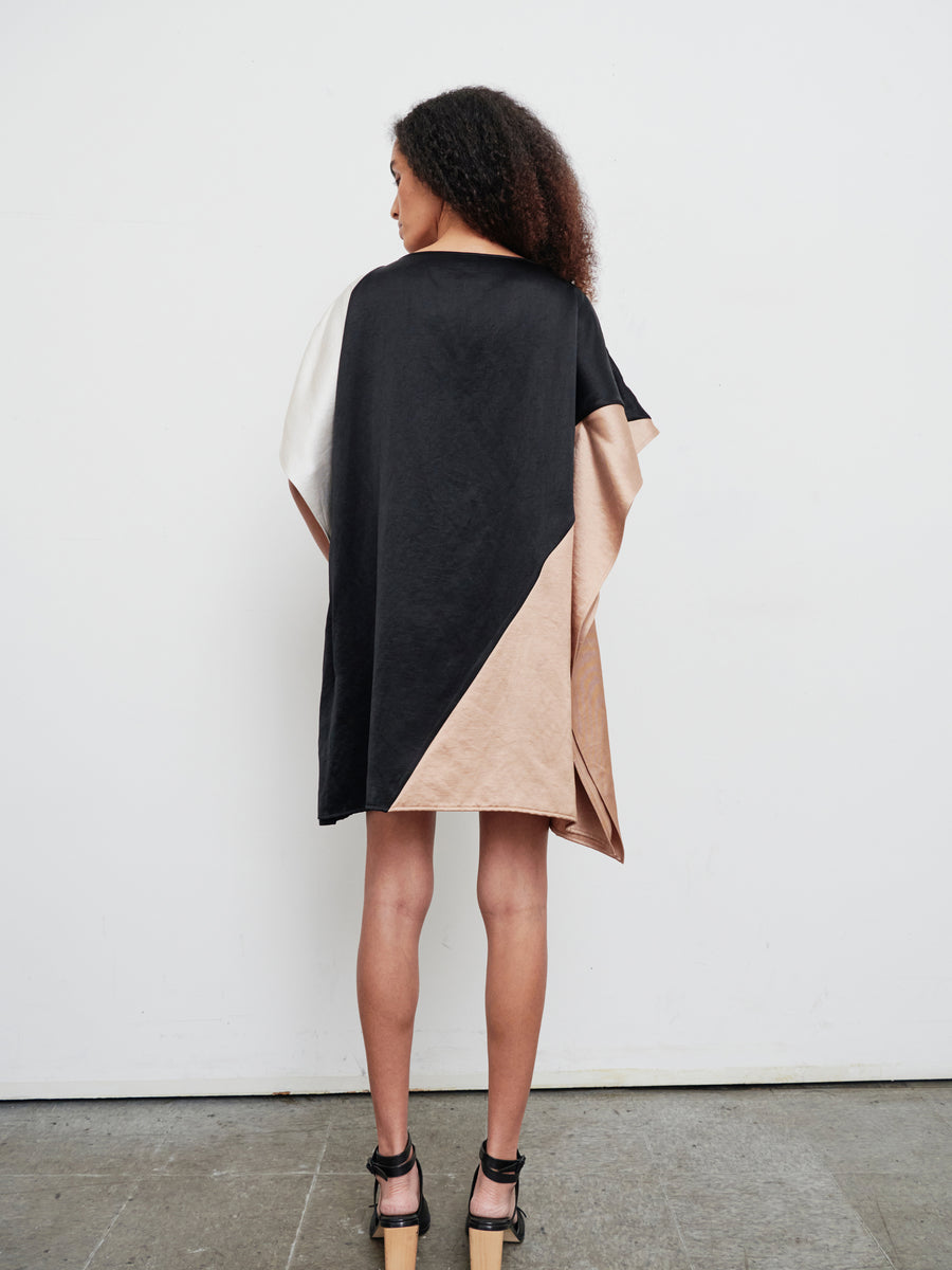 A person with curly hair is seen from the back, highlighting the knee-length Dia Dress by Zero + Maria Cornejo. The dress, made from a linen blend, features a striking color-block design in black, beige, and white with geometric shapes and falls just above the knees. They pair it with high-heeled sandals in a minimalist setting.