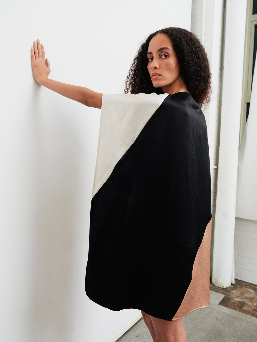 A person stands against a white wall, wearing the knee-length Dia Dress by Zero + Maria Cornejo, featuring a cape-style design with sections in black, white, and tan. They look over their shoulder with curly hair in a softly illuminated minimalist setting enhanced by natural light.