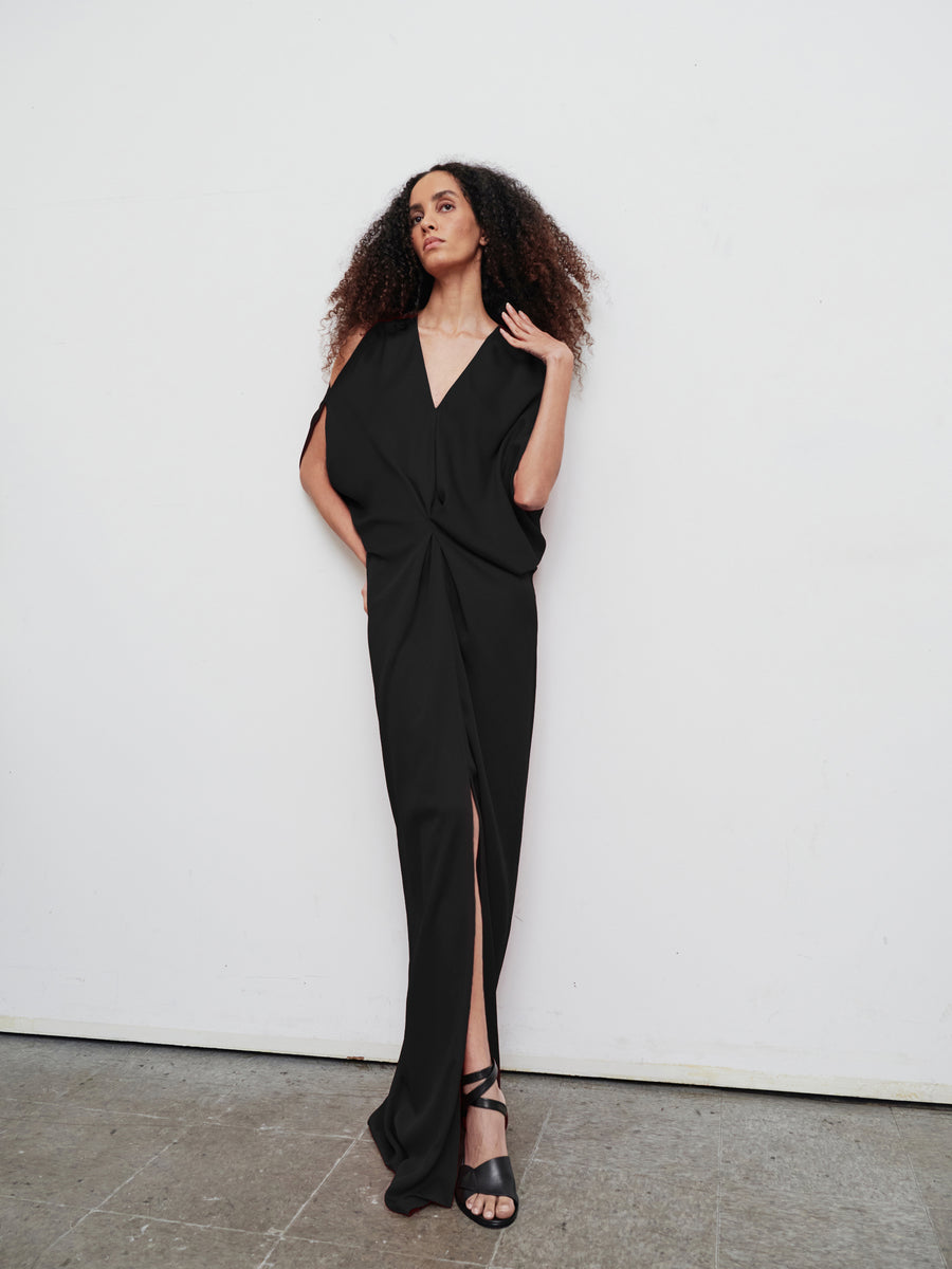 A woman stands against a plain white wall wearing the stylish Long Miu Dress by Zero + Maria Cornejo. This woven viscose dress features a deep V-neck and front slit. Her curly hair cascades over her shoulders, and she completes the look with black sandals, embracing an eco drape made from sustainably-sourced materials.