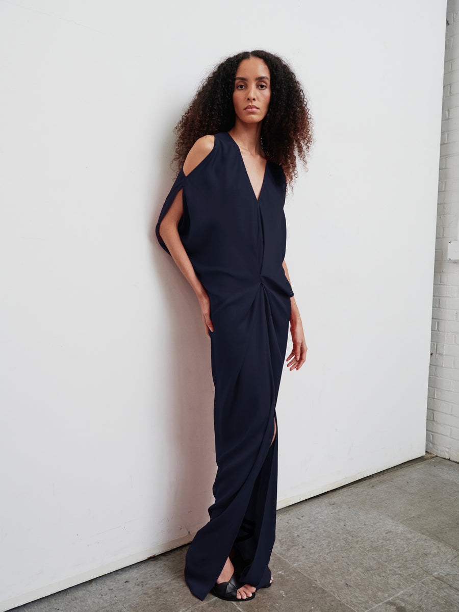 A person with curly hair wears the Long Miu Dress by Zero + Maria Cornejo, a navy floor-length piece crafted from woven viscose and featuring chic shoulder cutouts. The sustainably-sourced fabric of the dress drapes elegantly to the floor, showcasing a front slit as they stand against a white wall with a neutral expression.