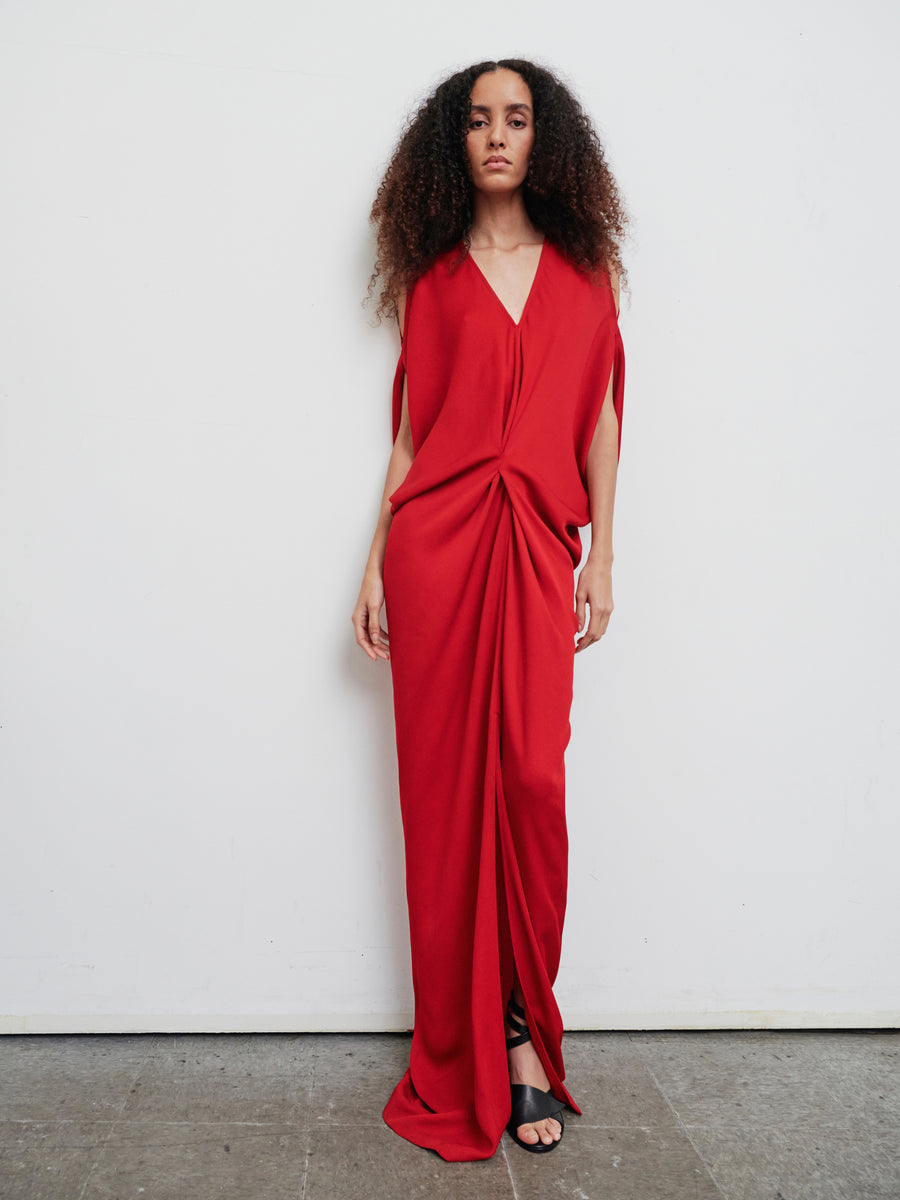 A woman stands against a plain wall dressed in the Long Miu Dress by Zero + Maria Cornejo. This elegant red dress, made from woven viscose with eco drape details, complements her curly hair as she poses with one hand by her side. Her expression is neutral, and she completes the look with black sandals.