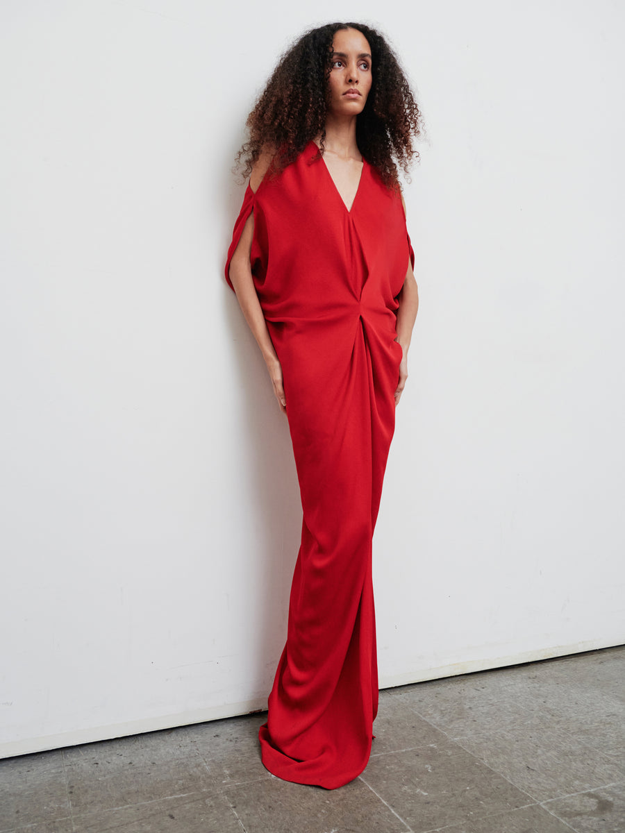 A person with curly hair is wearing the Long Miu Dress by Zero + Maria Cornejo—a flowing red woven viscose piece made from sustainably sourced materials. The dress features a loose fit with draped, gathered details. Standing against a plain white wall, their hands in pockets convey a relaxed and confident pose.