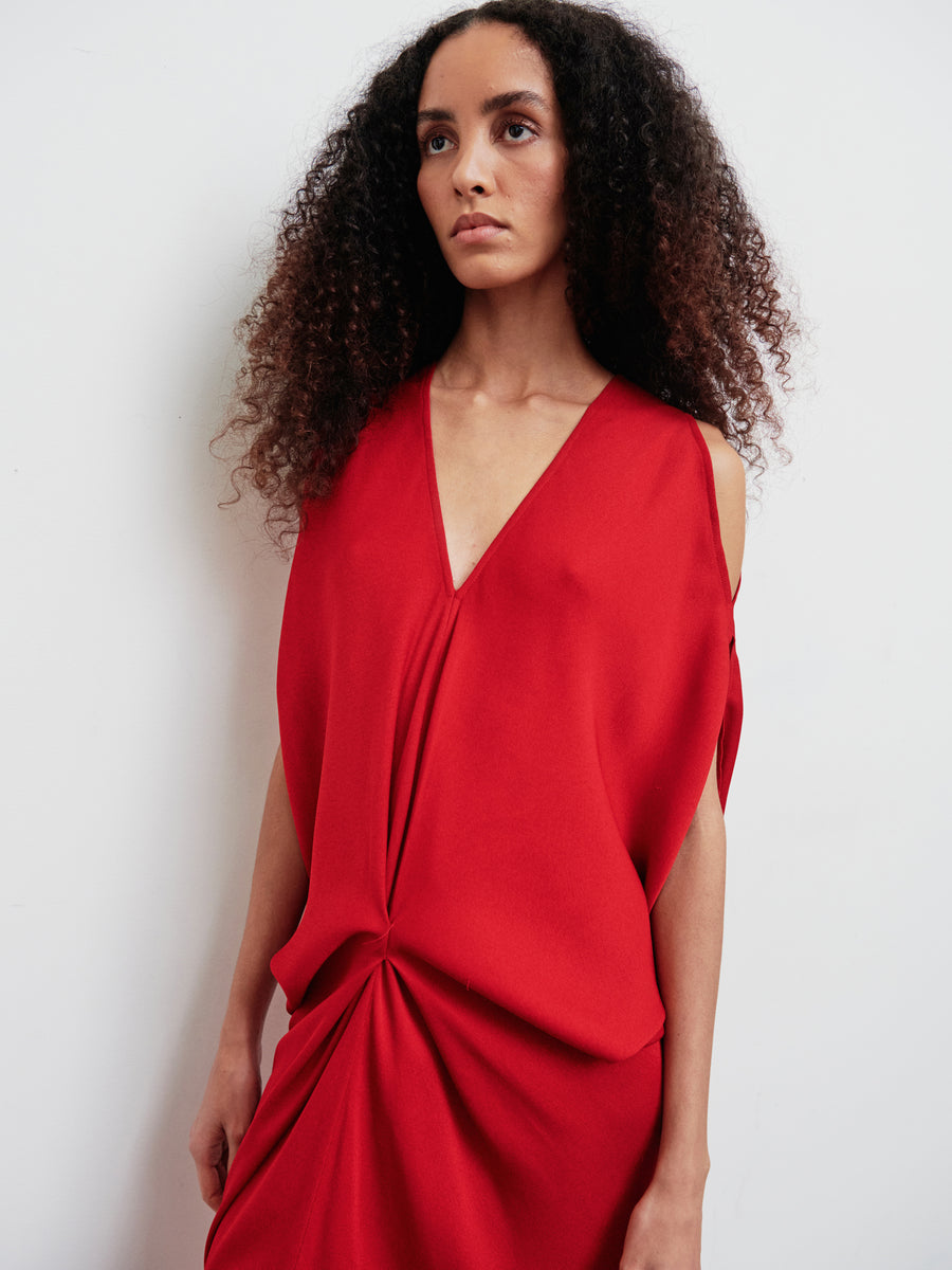 A person with long, curly brown hair is wearing the Long Miu Dress by Zero + Maria Cornejo. This sleeveless red dress, crafted from sustainably-sourced woven viscose, features an elegant eco drape and a twist detail at the waist. Set against a plain white background, they gaze slightly to the side.