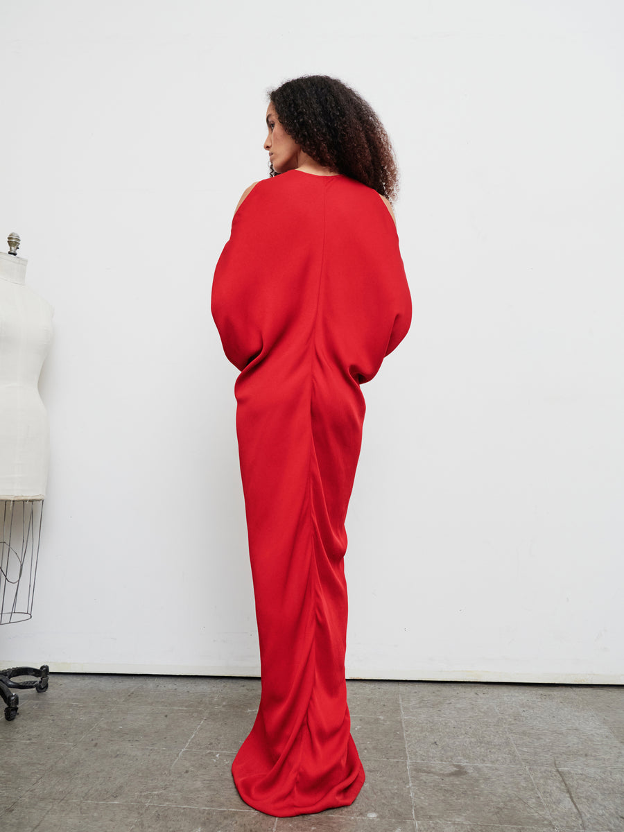 A person stands in the Long Miu Dress by Zero + Maria Cornejo, facing away from the camera. The sustainably-sourced, red woven viscose dress features a flowing design and dramatic sleeves. They stand on a gray concrete floor, near a dress form to the left.