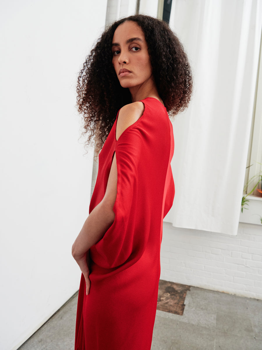 A woman with curly hair is wearing the Long Miu Dress by Zero + Maria Cornejo. The dress features a red color and an open-shoulder design crafted from woven viscose. She stands sideways in a minimalist room with white walls and an eco drape curtain, glancing over her shoulder toward the camera.