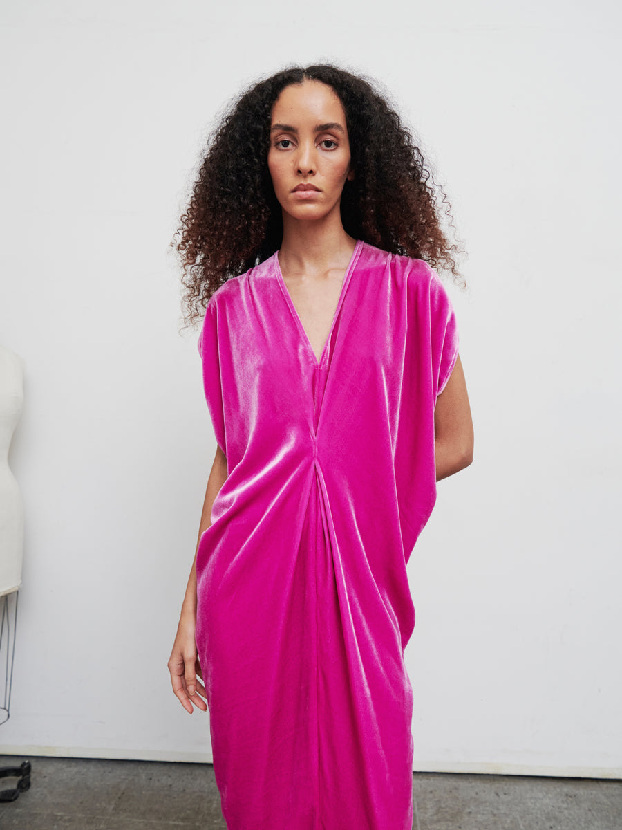 A person with curly hair is wearing the Long Issa Dress from Zero + Maria Cornejo, a vibrant pink velvet piece with cap sleeves, standing against a plain white background. A dress form is partially visible on the left side of the image.