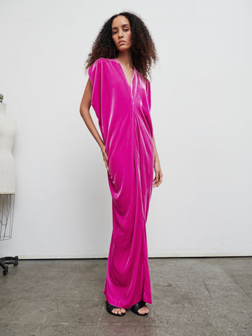 In a studio setting, an individual with curly hair models the Long Issa Dress by Zero + Maria Cornejo, a vibrant pink velvet dress featuring cap sleeves. A dress form can be seen partially in the background on the left, with concrete flooring and plain white walls completing the scene.