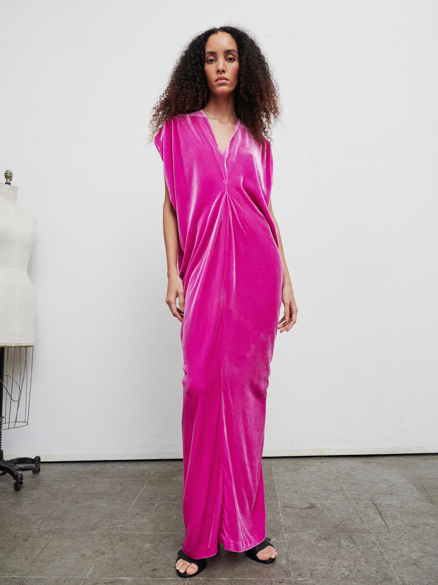 A person with curly hair is wearing the Long Issa Dress by Zero + Maria Cornejo, a long, bright pink velvet dress featuring cap sleeves, paired with black sandals. They stand on a concrete floor against a plain white wall, with a dress form partially visible in the background.