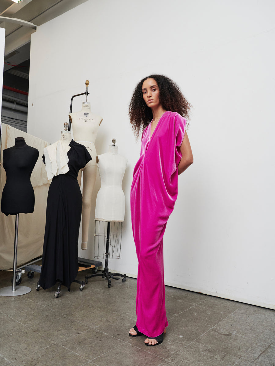 In the fashion studio, an individual with curly hair models a bright pink Long Issa Dress by Zero + Maria Cornejo. Behind them, a dress form features a velvet dress with cap sleeves, while another displays a partially draped white gown, all within the minimalist design of the room.