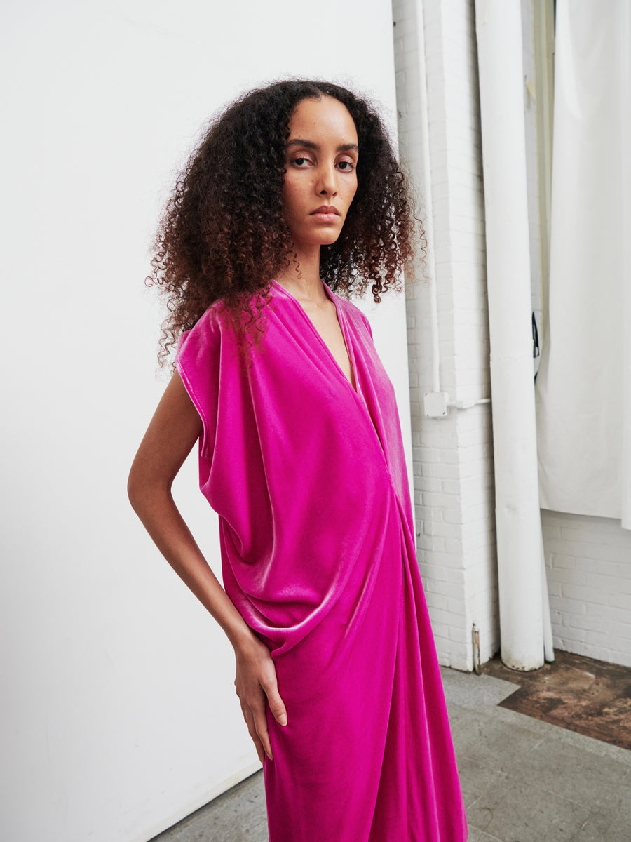 A person with curly hair is wearing the vibrant Long Issa Dress by Zero + Maria Cornejo, which showcases cap sleeves and a subtle back slit, as they stand in front of a white wall and brick pillar in a well-lit room. They have a calm expression, with one hand resting on their hip.