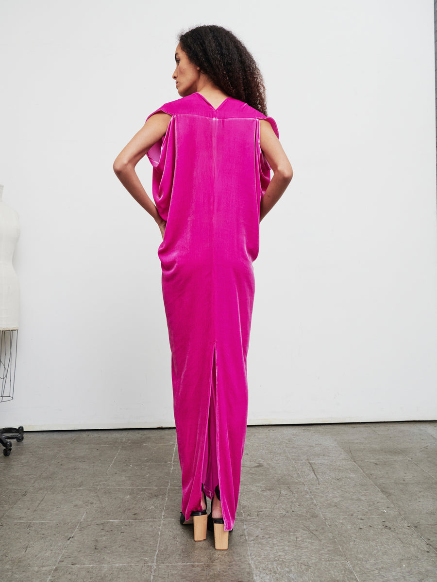 A person with long, curly hair is wearing the Long Issa Dress by Zero + Maria Cornejo, a vibrant velvet garment featuring cap sleeves and a deep V-back. They stand gracefully with their back to the camera in a minimalist room that features a dress form in the corner.