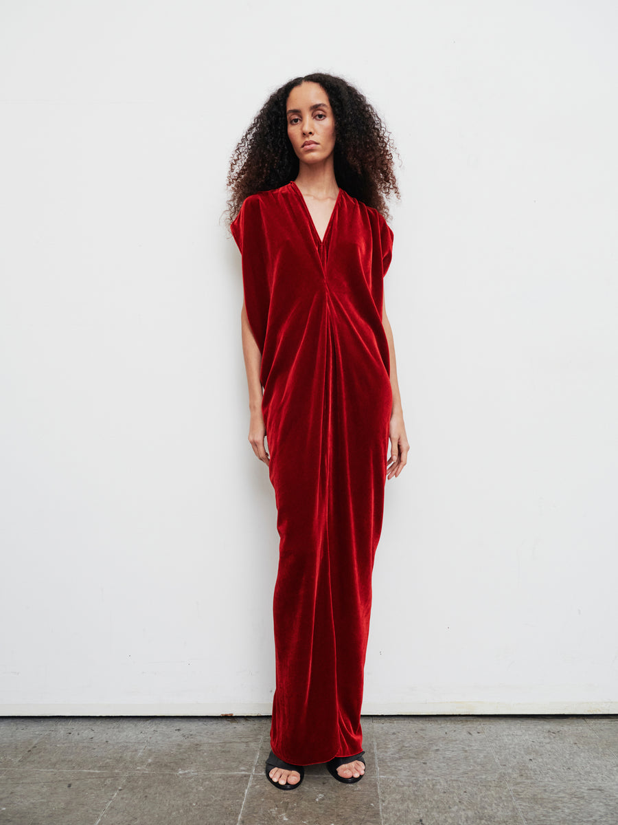 A person stands against a plain white background wearing the Long Issa Dress by Zero + Maria Cornejo, which is an elegant red velvet dress featuring a v-neck and cap sleeves. They have long curly hair and are wearing black sandals.