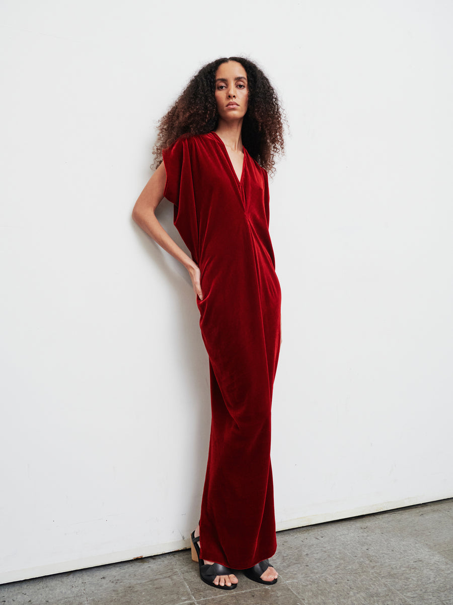 A person with long, curly hair stands against a white wall, elegantly dressed in the floor-length Long Issa Dress by Zero + Maria Cornejo, crafted from deep red velvet and featuring cap sleeves. They complete their sophisticated look with black open-toe heels. The minimalistic background accentuates the elegance of their attire.