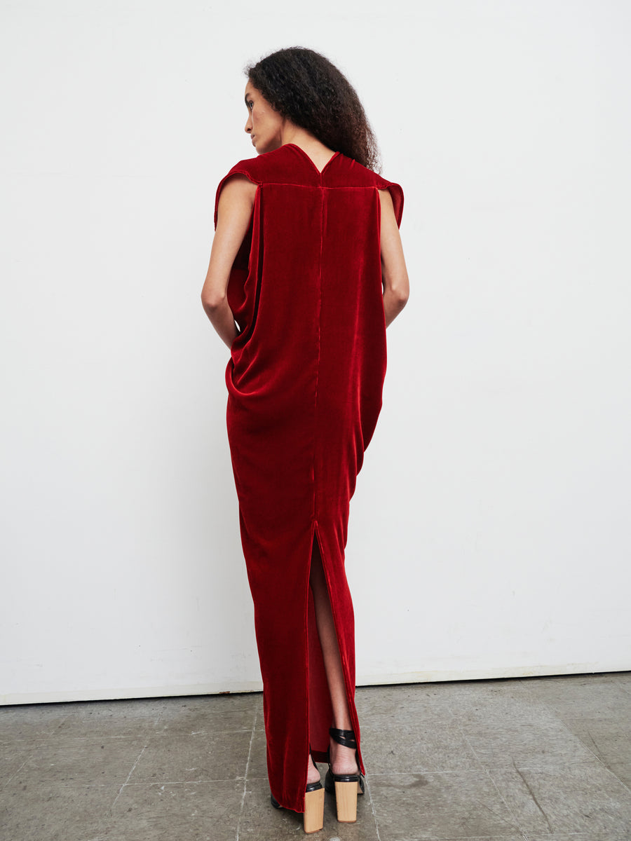 A person with long curly hair is wearing the Long Issa Dress by Zero + Maria Cornejo, which is a sleeveless, luxurious velvet dress in deep red and features a slit at the back. They stand on a concrete floor facing a white wall, complemented by black platform sandals.