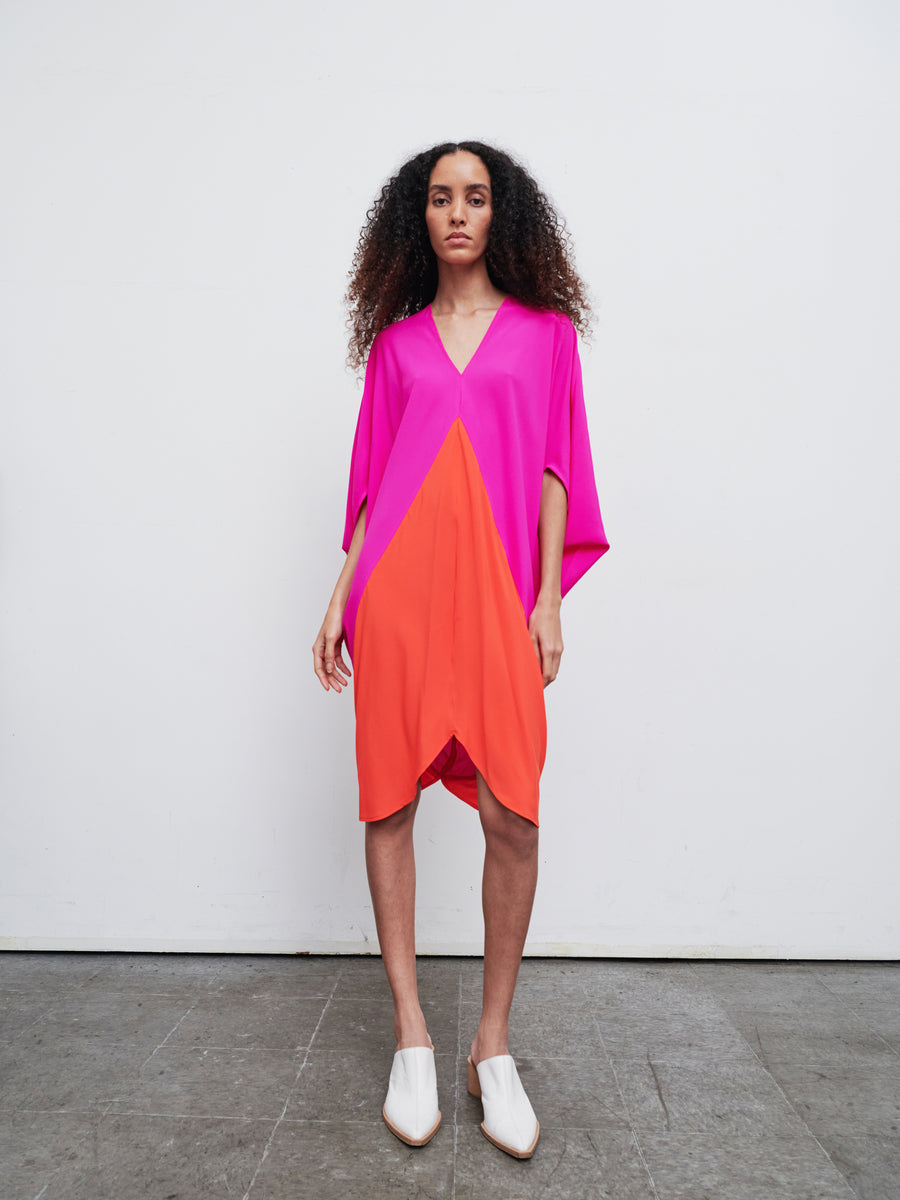 A person stands against a plain background wearing the Koya Dress by Zero + Maria Cornejo, featuring a vibrant cocoon silhouette with bright pink and orange panels. They have long curly hair and are paired with white shoes.