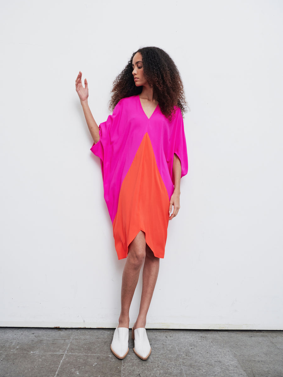 A person with curly hair is seen wearing the vibrant Koya Dress from Zero + Maria Cornejo, which features pink and orange sections. The dress's cocoon silhouette perfectly enhances their style as they pose against a white background, one arm slightly raised, while elegantly donning white pointed shoes.