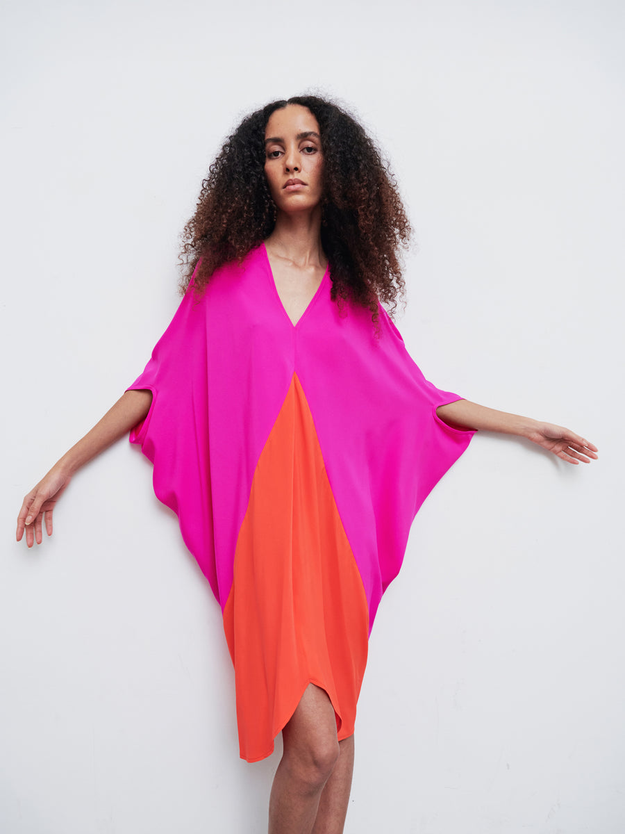 A woman with curly hair poses against a white background, wearing the Koya Dress by Zero + Maria Cornejo. The dress is made of brightly colored silk charmeuse with a pink top and orange bottom, featuring wide sleeves and a loose fit. The cocoon silhouette accentuates her form as her arms extend outward.