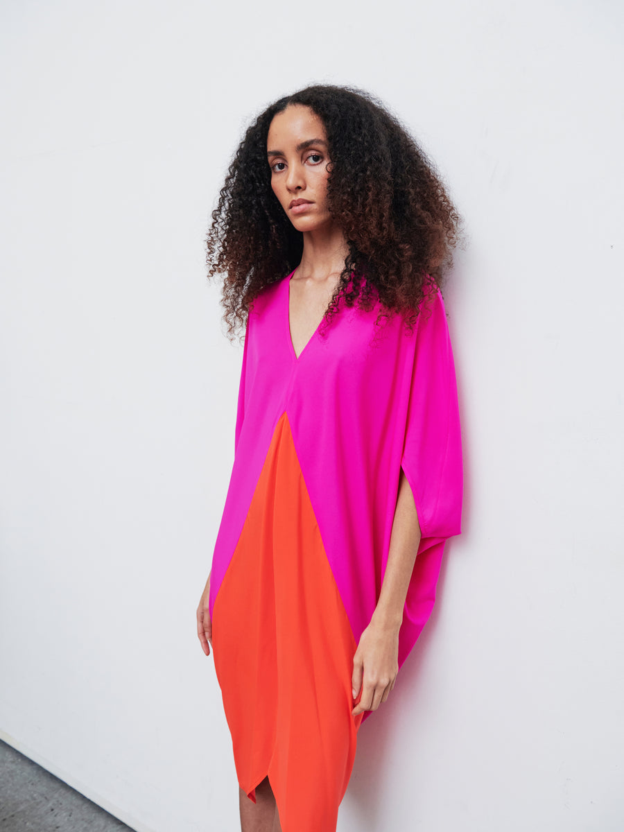 A person with curly hair, wearing the Koya Dress by Zero + Maria Cornejo in a vibrant silk charmeuse cocoon silhouette, leans against a white wall. The dress elegantly transitions from a pink v-neck top to an orange bottom.