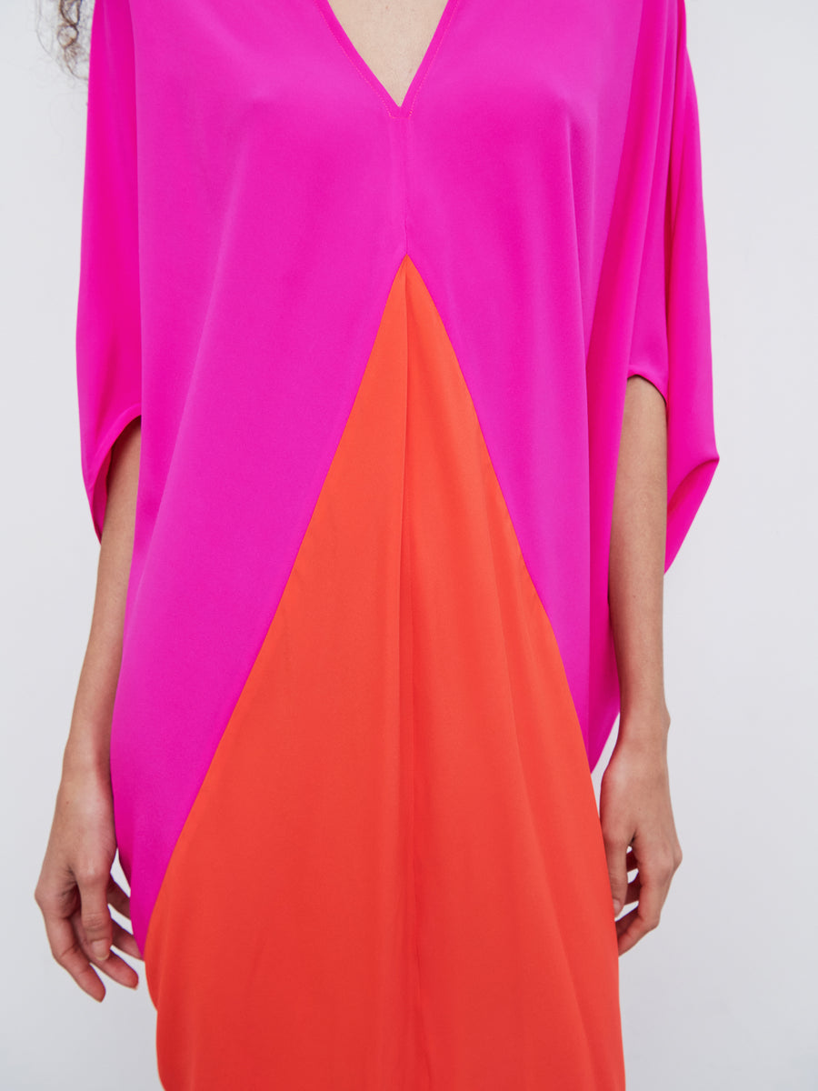 A person is wearing the Koya Dress by Zero + Maria Cornejo, a vibrant silk charmeuse piece with a pink top section and an orange triangular bottom section. This V-neck dress, with its short sleeves, showcases an elegant cocoon silhouette. The image highlights the upper body.