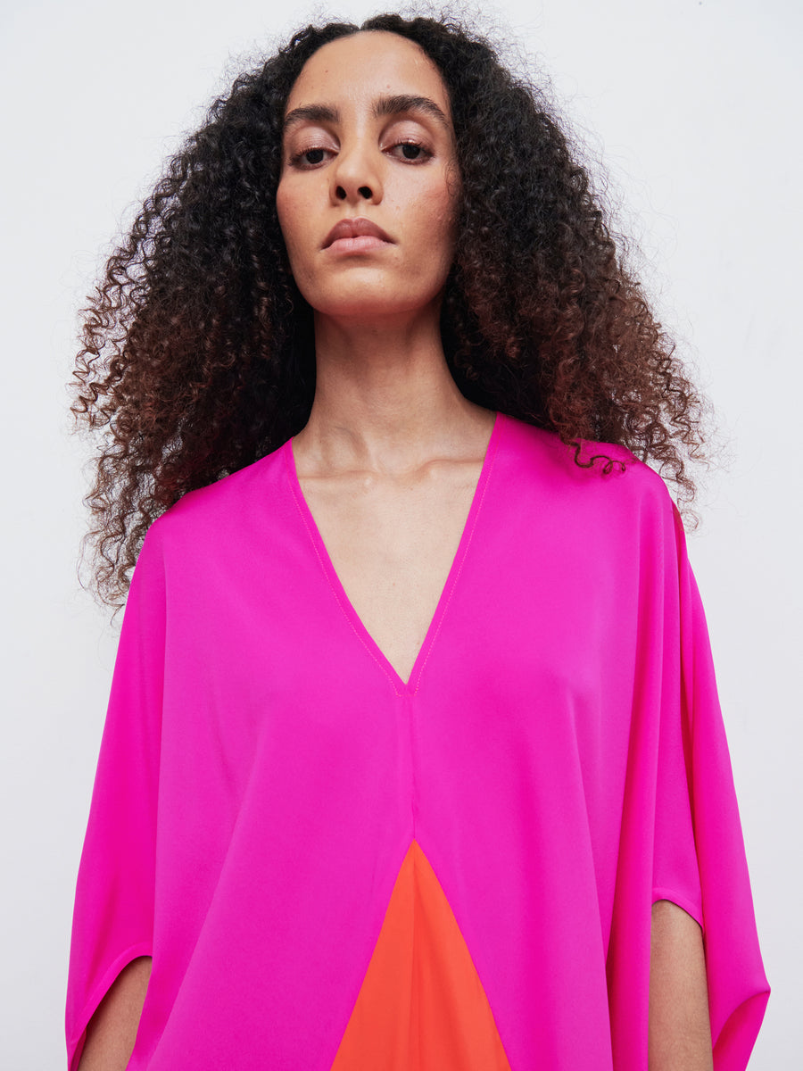 A person with curly hair dons the vibrant pink Koya Dress by Zero + Maria Cornejo, which showcases an orange section and a cocoon silhouette as they gaze directly at the camera against a plain white background.