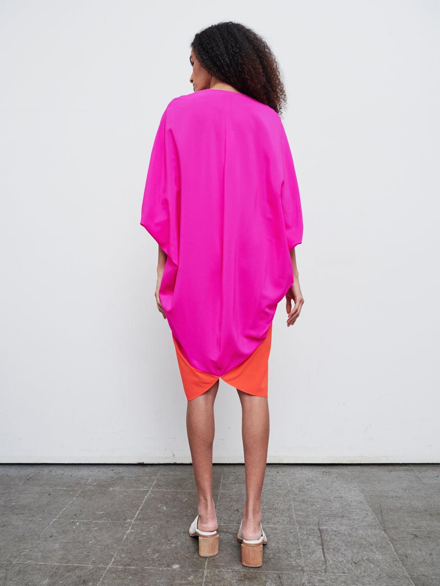 A person with curly hair stands with their back to the camera, wearing a bright pink and orange Koya Dress by Zero + Maria Cornejo. The dress, resembling a cocoon silhouette, contrasts the concrete floor and plain white background.