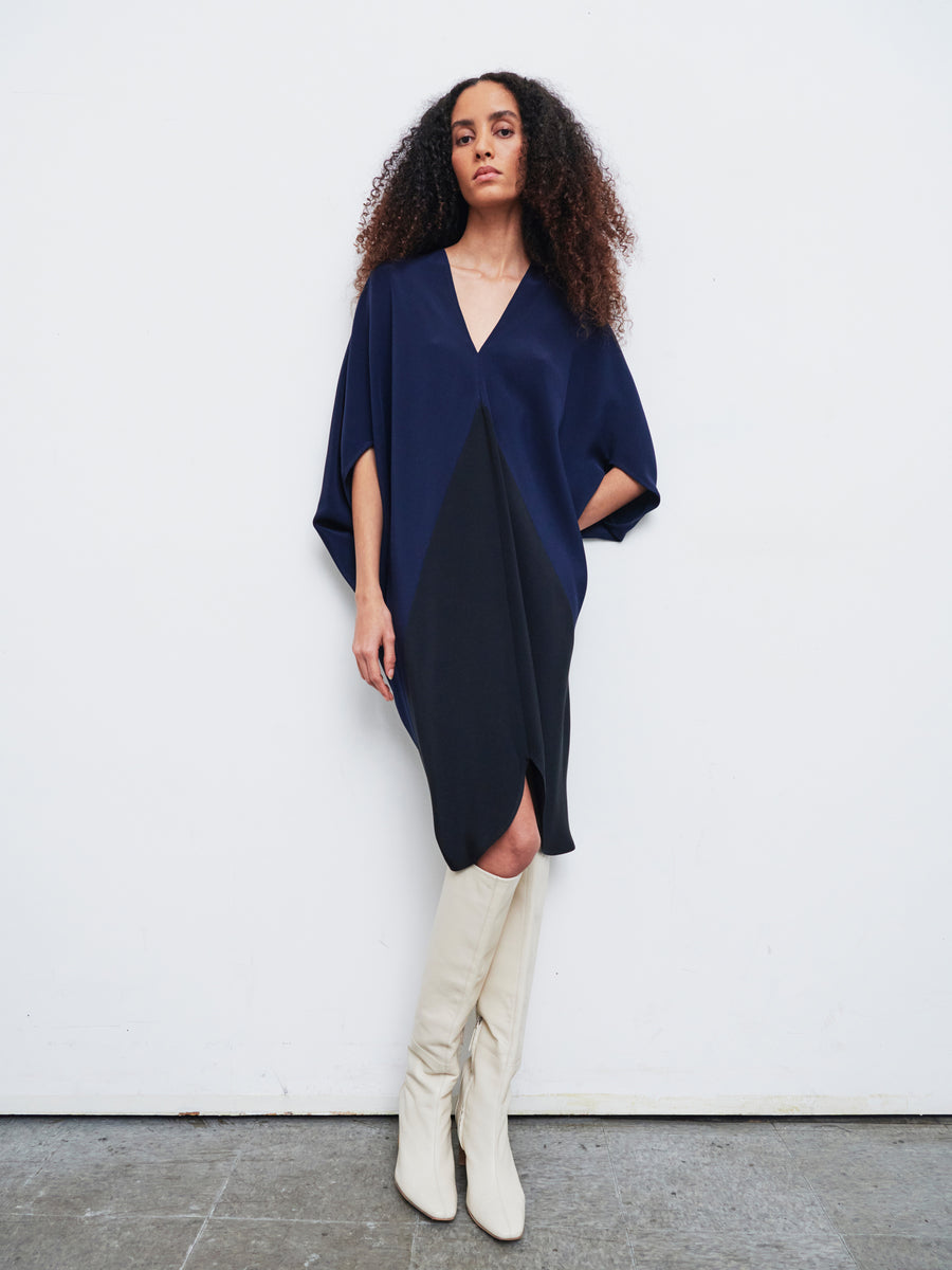 Standing against a white wall, a person with long, curly hair models the Koya Dress by Zero + Maria Cornejo. This dual-tone navy and black silk crepe V-neck piece has elegant batwing sleeves, and its cocoon silhouette features convenient pockets where they casually repose their hands. The look is perfectly completed with tall white boots.