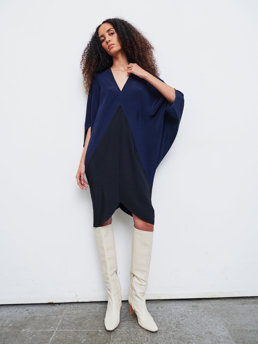 A person with curly hair poses confidently against a plain background, wearing the Koya Dress by Zero + Maria Cornejo. This dark blue silk crepe dress has an asymmetrical design and is paired with tall white boots. Its striking geometric panel in black accentuates the elegant cocoon silhouette.