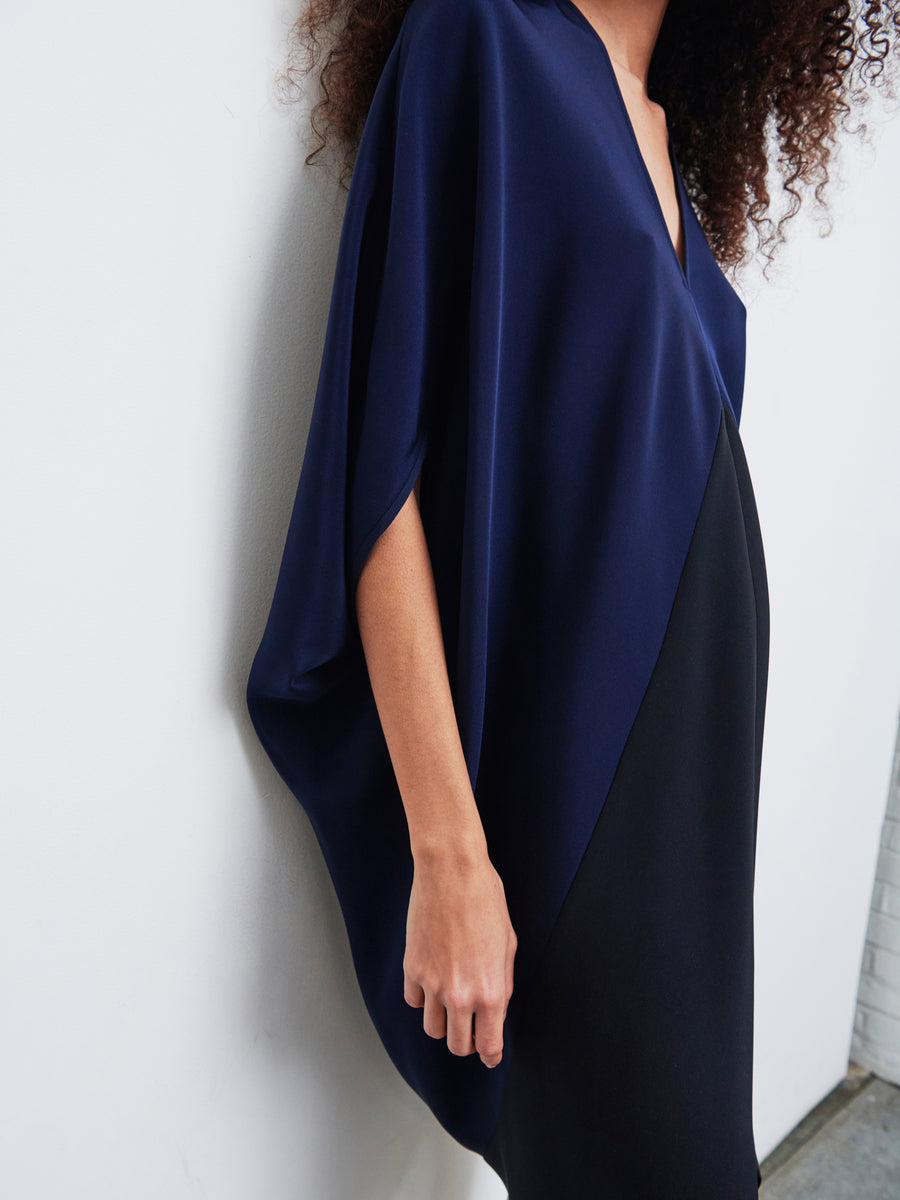 A person wearing the Koya Dress by Zero + Maria Cornejo, a dark blue, loose-fitting top with a deep v-neckline, stands against a white wall. Featuring flowing sleeves and a minimalist design, the elegant dress showcases a hint of cocoon silhouette. The person's curly hair is partially visible.