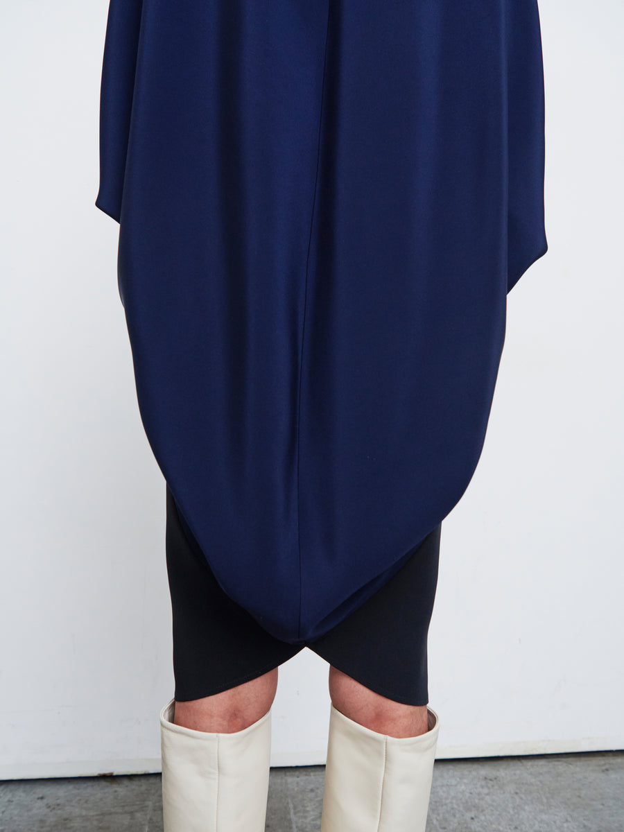 A person wearing the Koya Dress by Zero + Maria Cornejo, featuring a dark navy blue silk crepe with draped detailing over a black skirt, paired with tall white boots, stands against a light background.