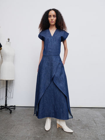 A person with long, curly hair stands on a concrete floor wearing the Mido Dress by Zero + Maria Cornejo, crafted from GOTS-certified cotton. The outfit includes a short-sleeved top and a long, layered skirt. With one hand in their pocket, they pair the look with white boots. A mannequin and dress form can be seen in the background.