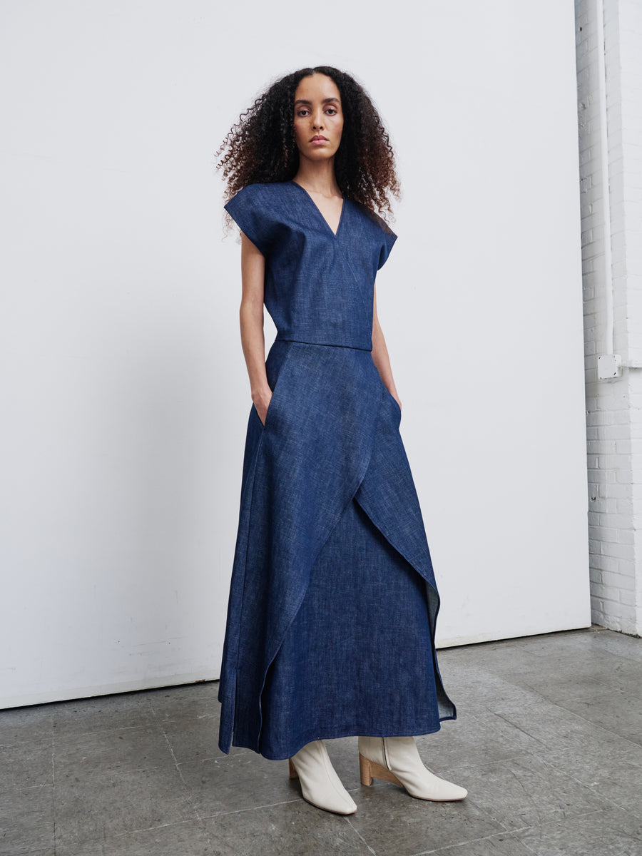 A person with curly hair wears the Mido Dress by Zero + Maria Cornejo, a fashionable ensemble crafted from organic denim, showcasing a sleeveless V-neck top and a long, layered skirt. They stand in a minimalist room with a light background and concrete floor, paired with white heeled boots.