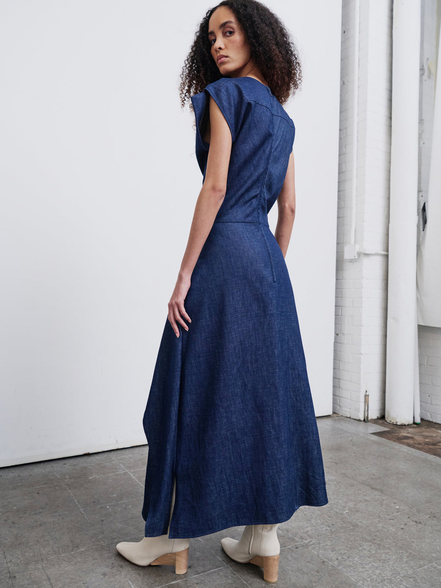 A person with curly hair wearing a sleeveless long blue Mido Dress from Zero + Maria Cornejo, paired with white ankle boots, stands in an industrial-style room, gazing over their shoulder.