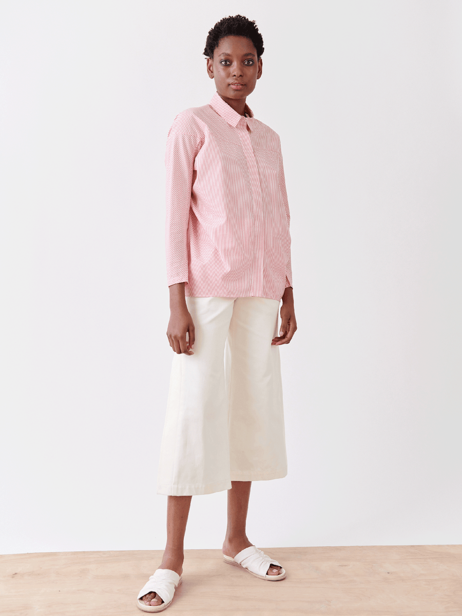 A person wearing the Manu Shirt by Zero + Maria Cornejo, featuring menswear-inspired pink and white stripes in cotton, pairs it with white wide-legged pants and slide sandals. They pose against a plain light background with a neutral expression, exuding effortless summer shirting style.