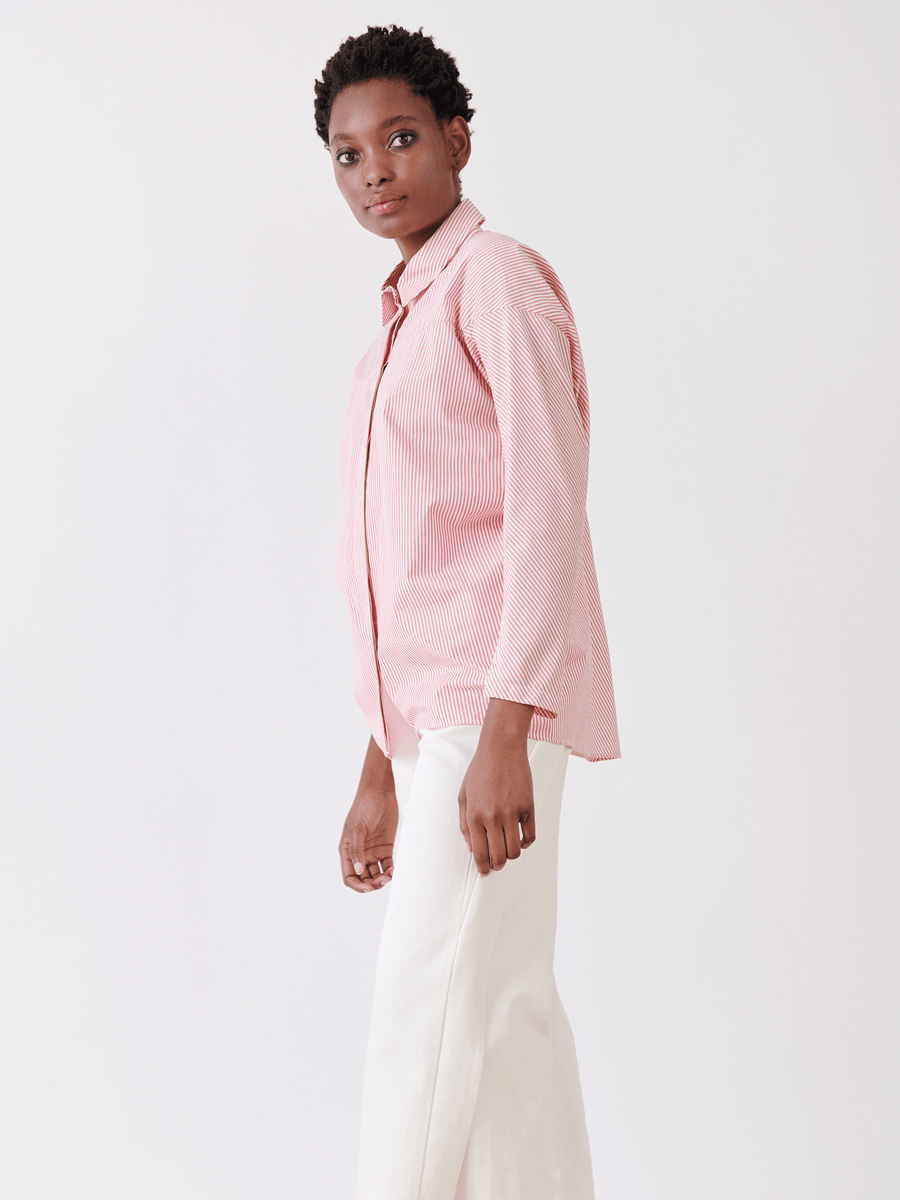 Against a light backdrop, a person in the Manu Shirt by Zero + Maria Cornejo, featuring menswear-inspired pink stripes and cotton fabric, pairs it with white pants. With short curly hair and a side glance, they effortlessly capture summer shirting style.