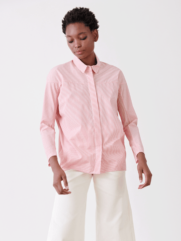 Dressed in a menswear-inspired Manu Shirt by Zero + Maria Cornejo, featuring pink and white stripes, a person stands against a plain white background. Paired with crisp white pants, their short hair and relaxed expression highlight the effortless summer style.