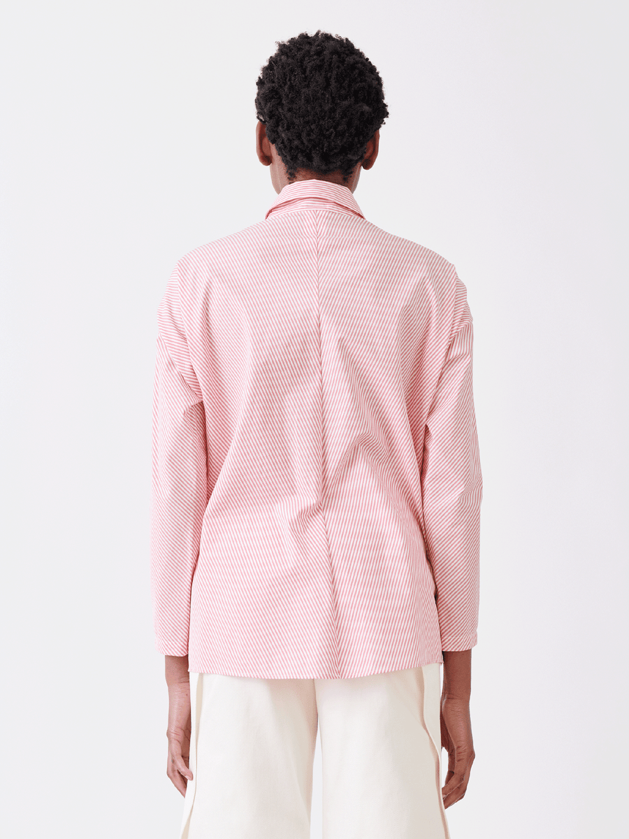 Facing away, a person with short curly hair dons the Manu Shirt—a menswear-inspired pink and white striped blazer by Zero + Maria Cornejo—and light pants, set against a plain white backdrop, capturing summer shirting vibes.