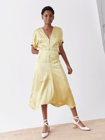 A person poses confidently with one hand on their hip, wearing the Aissa Joi Dress by Zero + Maria Cornejo. This linen-blend dress in light yellow features a V-neck and tiered skirt, complemented by white heels on a wooden floor against a white background.