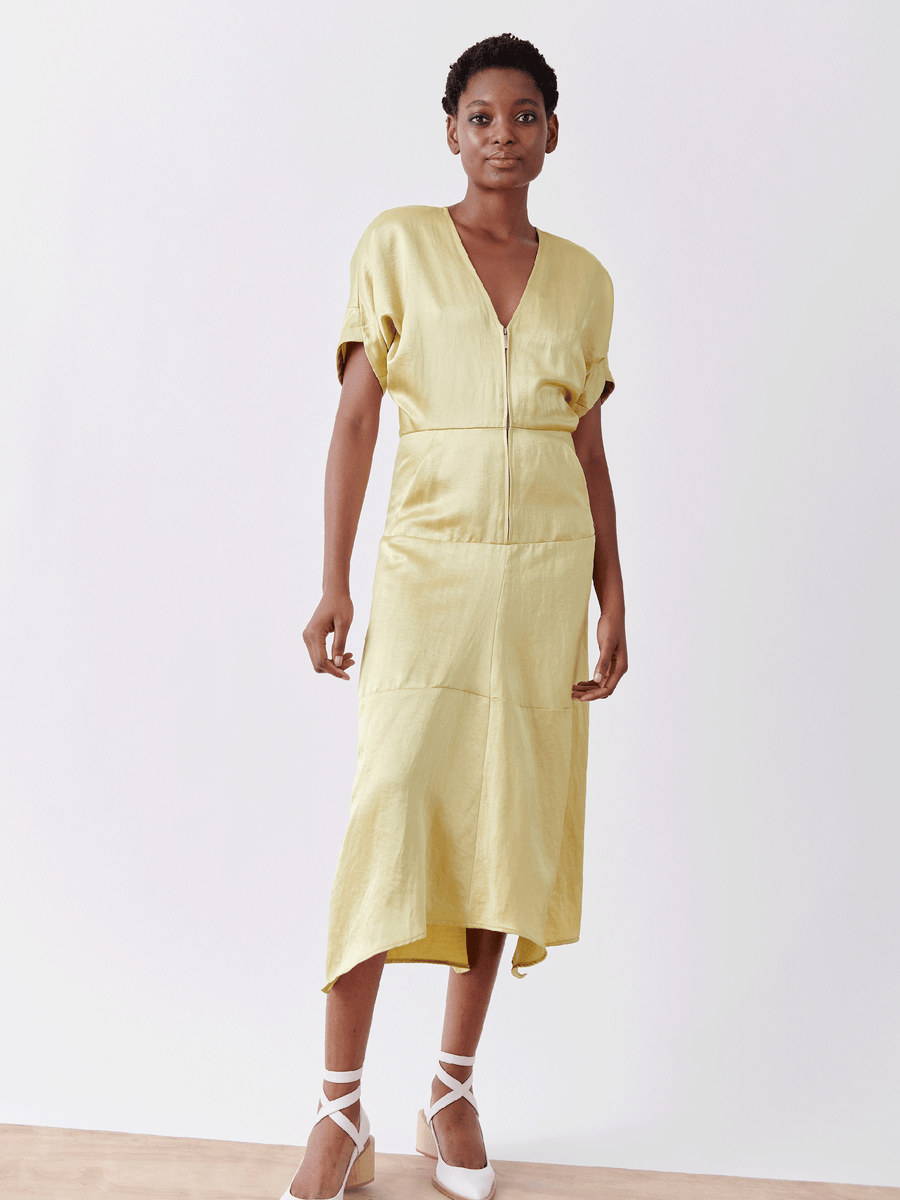 A person confidently wears the Aissa Joi Dress by Zero + Maria Cornejo, a light yellow, short-sleeved V-neck linen-blend with an asymmetrical hemline. They pair it elegantly with white strappy heels against a plain white background.