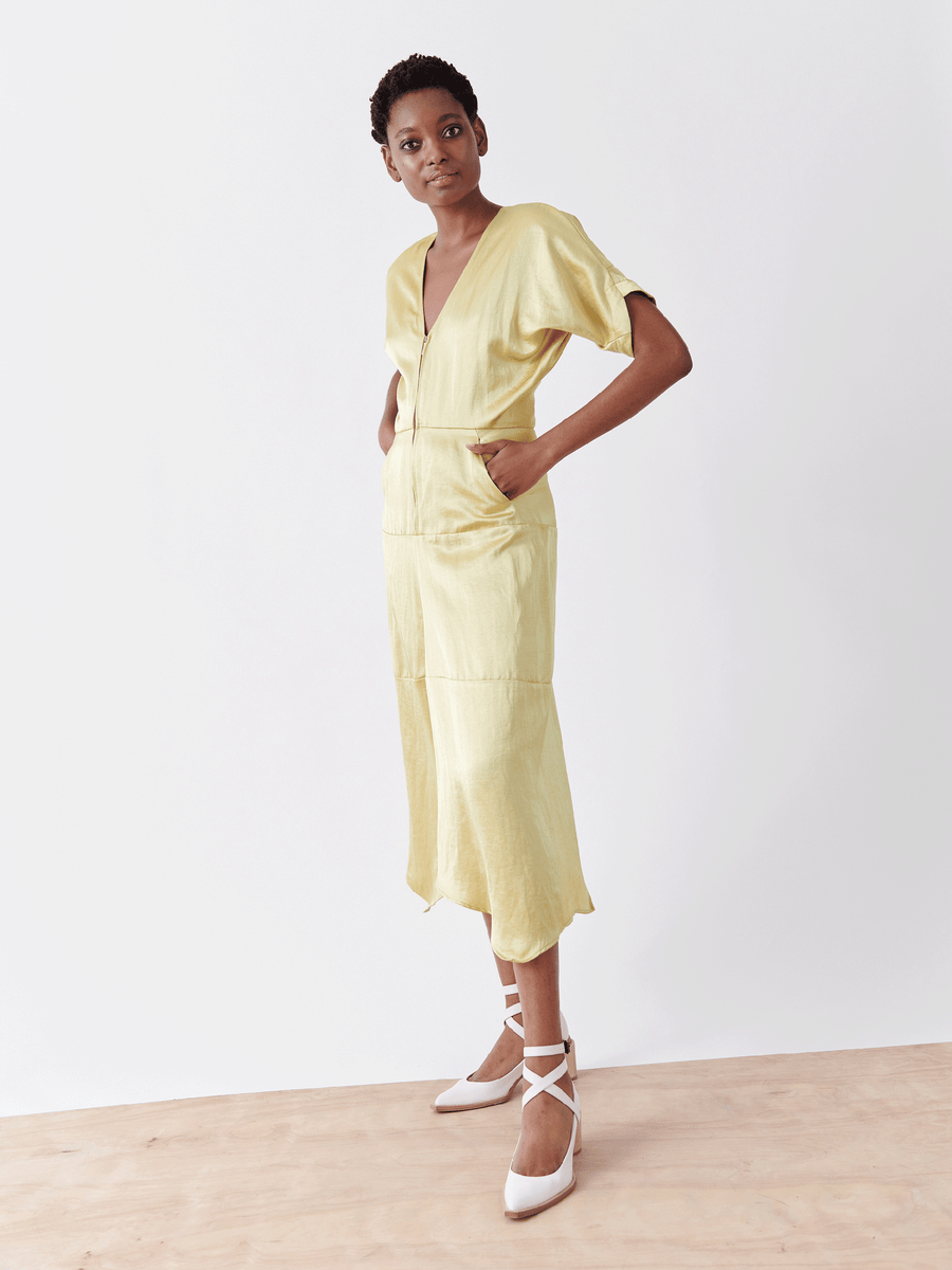 Dressed in Zero + Maria Cornejo's Aissa Joi Dress, a light yellow, knee-length linen-blend piece with a zippered front and pockets, the person stands on a wooden floor against a white backdrop. White high-heeled shoes with crisscross straps complete the look.