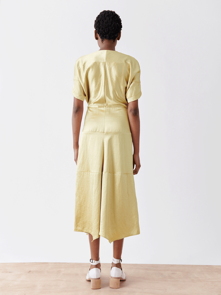 Rear view of a person with short hair wearing the Aissa Joi Dress from Zero + Maria Cornejo. The light yellow, linen-blend fabric looks breezy with white open-toe shoes on a wooden floor against a plain white background.