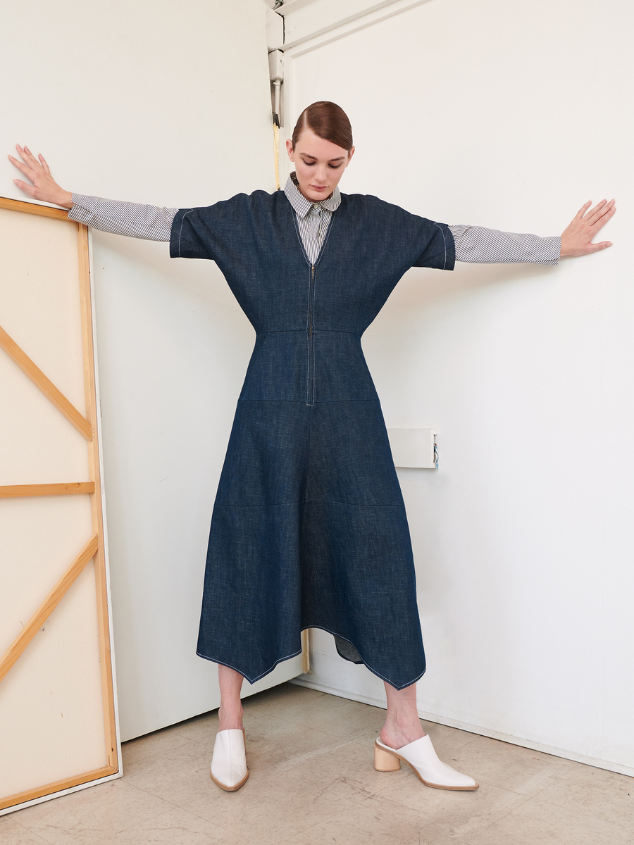 A person stands against a wall, arms outstretched, in an Aissa Joi Dress by Zero + Maria Cornejo layered over a long-sleeved striped shirt. They have short hair and sport white heeled mules. To the left, a framed canvas adds to their stylish look.