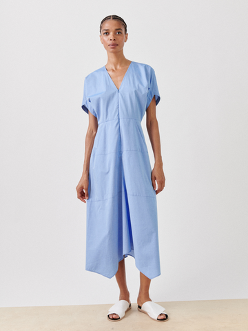 A person with a clean, minimalist appearance stands against a plain white background. They are wearing the Zero + Maria Cornejo Aissa Joi Dress, which is a light blue, V-neck, short-sleeved cotton chambray dress with a midi-length hem and white slip-on shoes. The dress has a relaxed fit and gently drapes over the body.