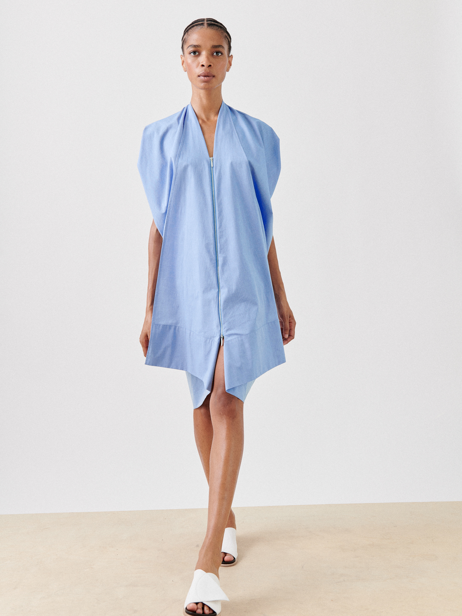 A woman is walking and wearing a light blue, zip-up, short-sleeve, cotton chambray Foil Dress by Zero + Maria Cornejo with an asymmetric hem. She also has on white, open-toe shoes. The background is plain white, and the floor appears beige.