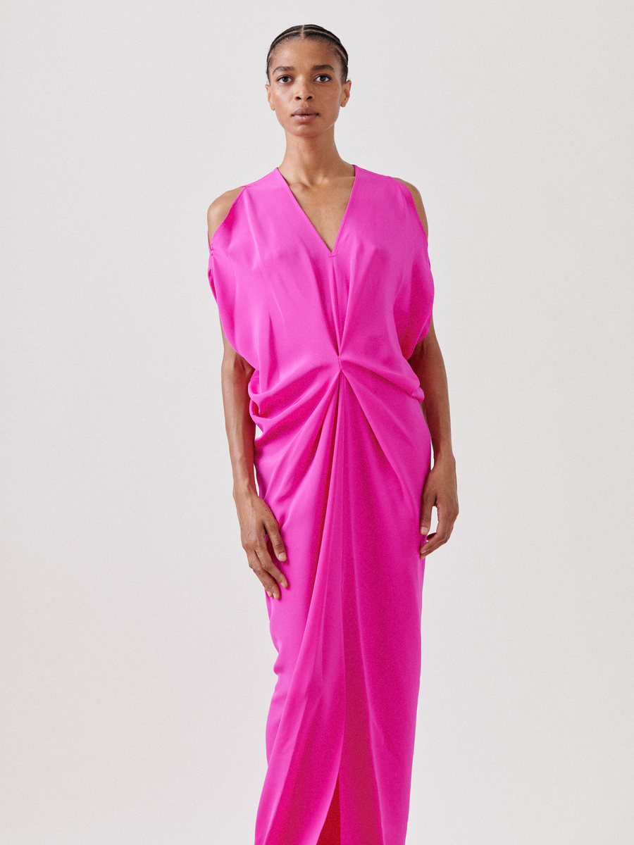 A person wearing the Long Miu Dress - Final Sale by Zero + Maria Cornejo, featuring vibrant pink silk, elegant cut-out shoulders, and a draped design, stands against a plain background. Their braided hair complements their neutral expression for a striking yet serene image.