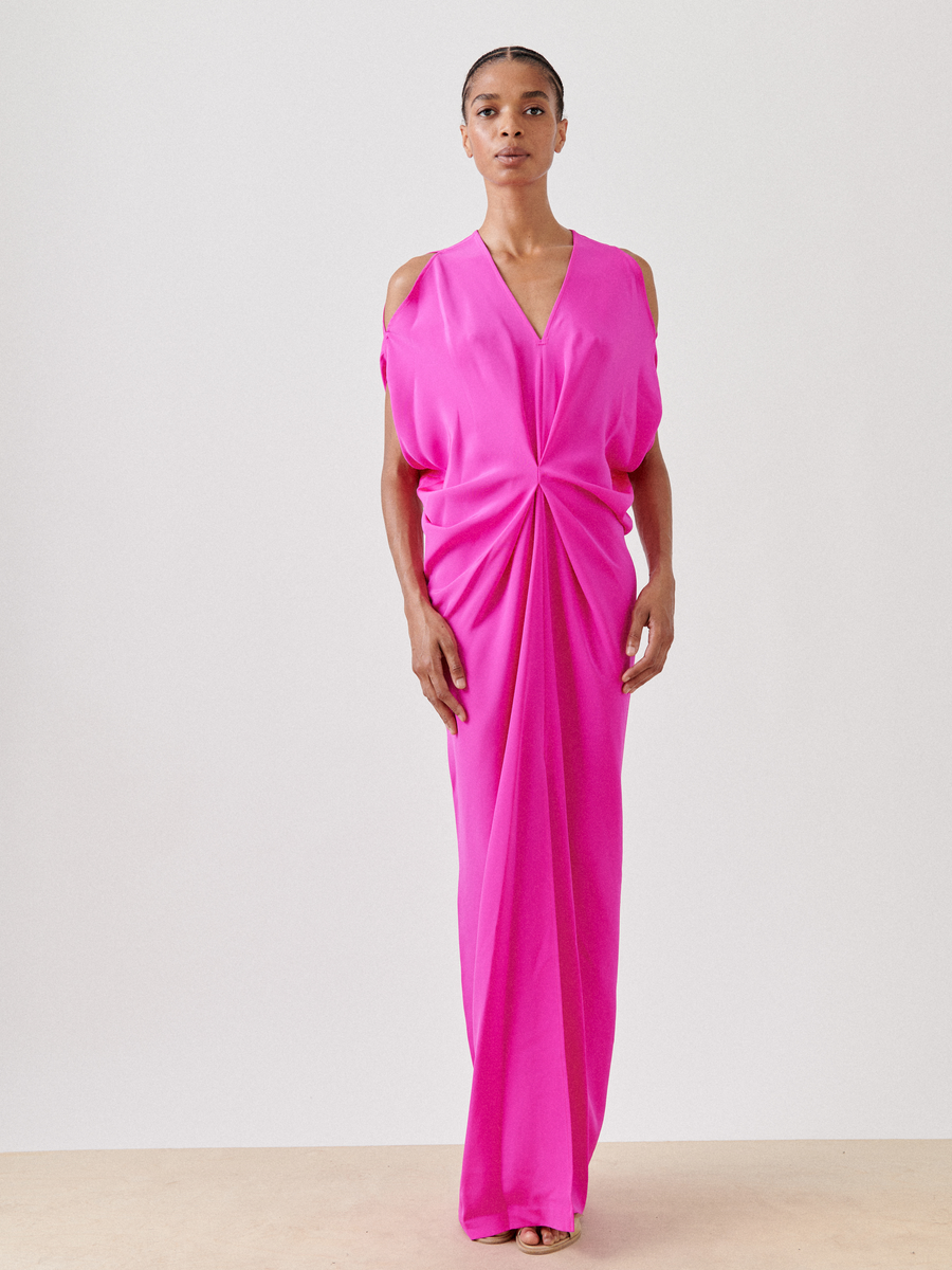 Against a plain background, a person wears the Long Miu Dress by Zero + Maria Cornejo in vivid pink silk, showcasing cut-out shoulders and a gathered twist design at the waist. The full-length dress flows elegantly as they gaze forward.